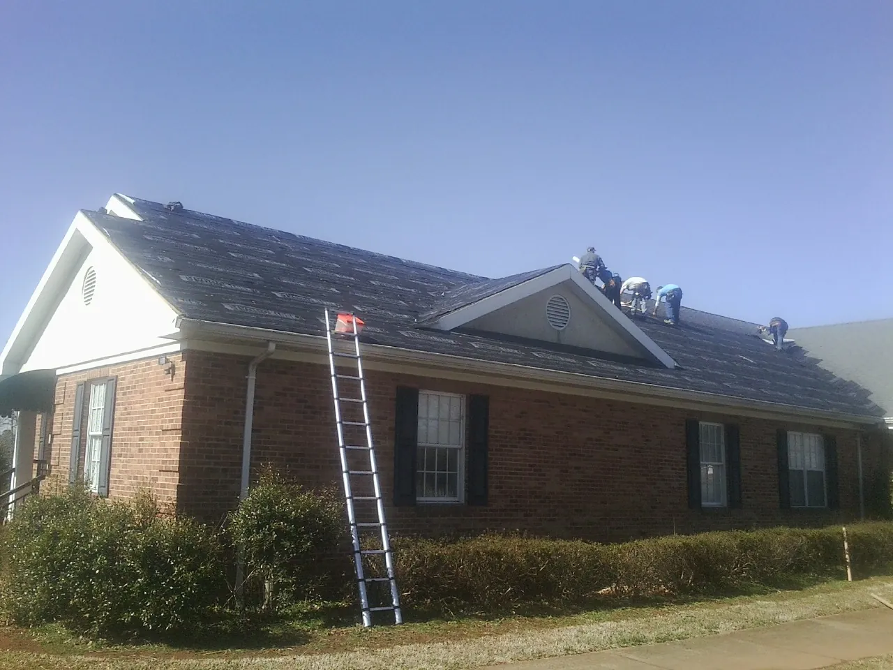 Crew tearing shingles off a brick ranch roof with a ladder set — Emergency Roof Repair work by our crew in Brockport