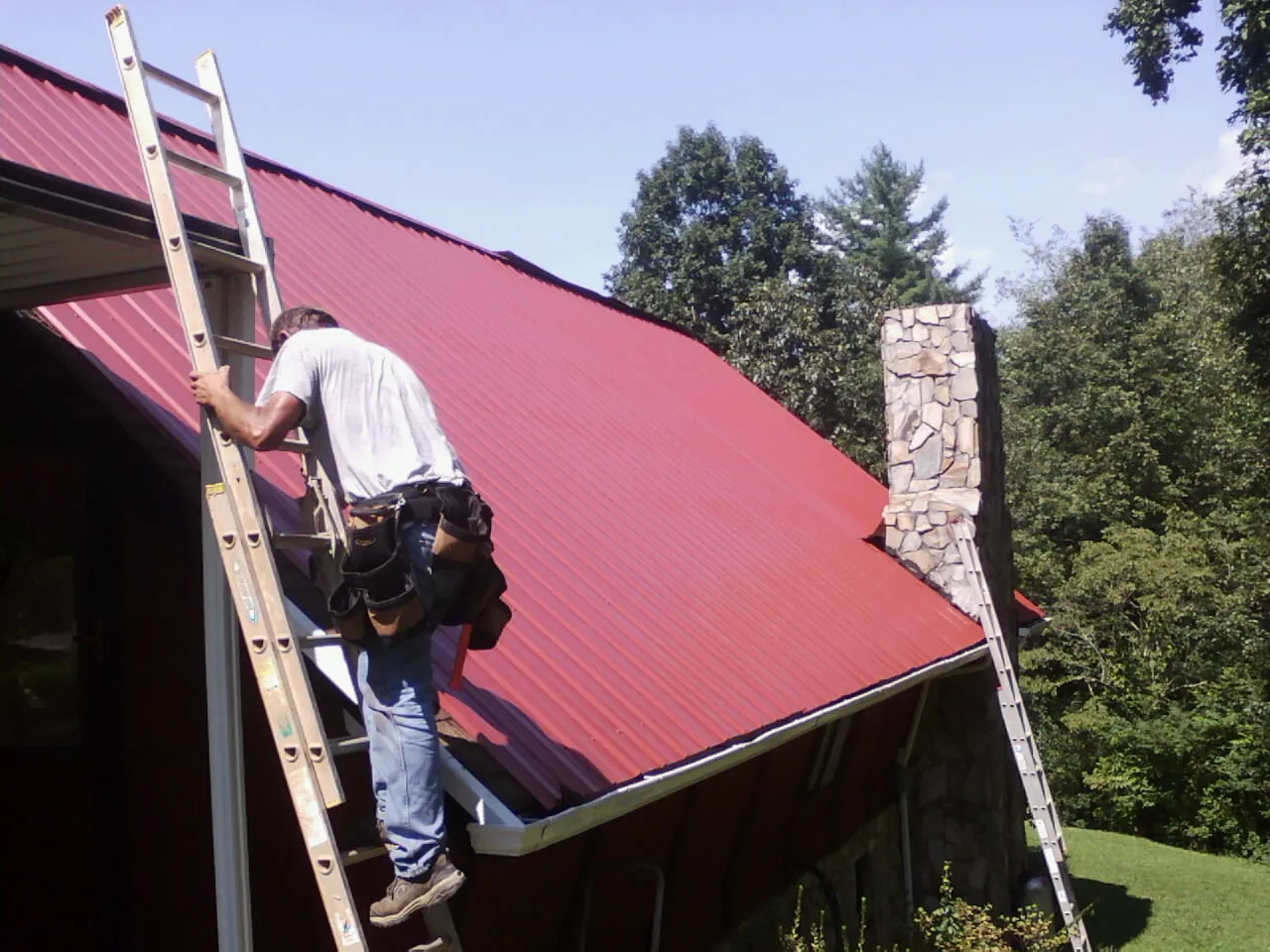 Roofer on a ladder beside a red metal roof and stone chimney — Soffit & Fascia Repair by Roofing Brothers in Brockport